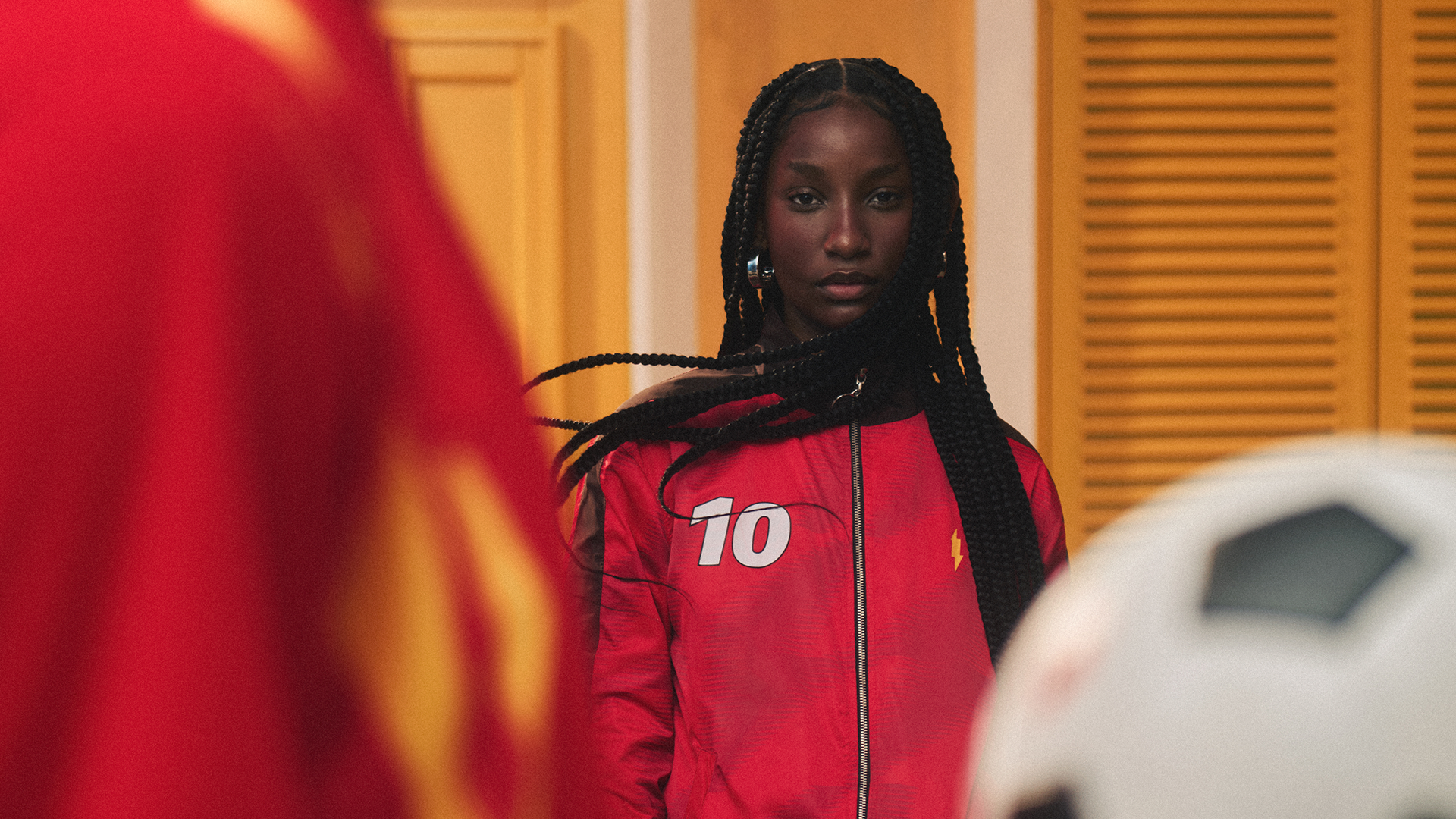 Jovem negra com tranças longas, vestindo jaqueta esportiva vermelha com o número 10 branco, olha séria para frente. Um ombro de outra pessoa em vermelho e uma bola de futebol branca e preta aparecem em primeiro plano, desfocados. Ao fundo, uma parede amarela com persianas