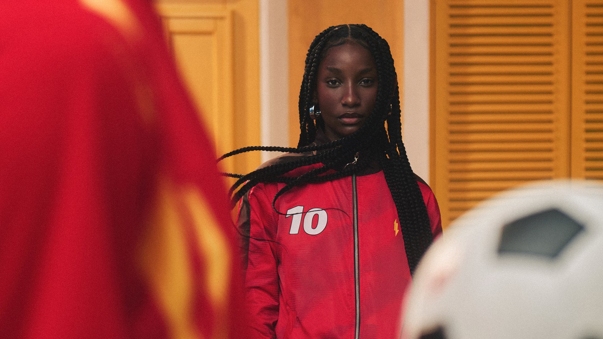 Jovem negra com tranças longas, vestindo jaqueta esportiva vermelha com o número 10 branco, olha séria para frente. Um ombro de outra pessoa em vermelho e uma bola de futebol branca e preta aparecem em primeiro plano, desfocados. Ao fundo, uma parede amarela com persianas