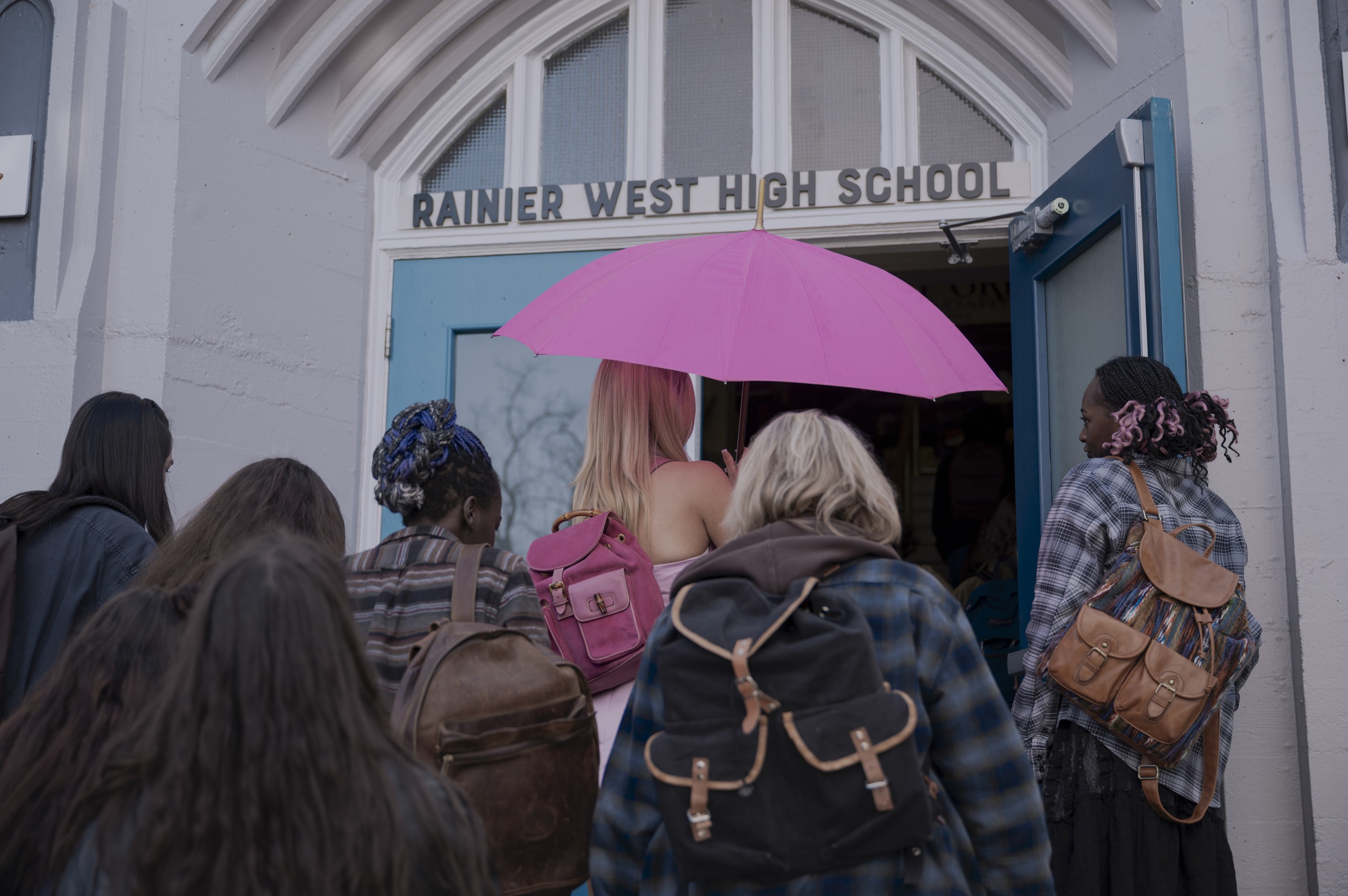 Grupo de estudantes, a maioria de costas, entrando na escola Rainier West High School. Uma garota loira com guarda-chuva rosa e mochila rosa se destaca no centro
