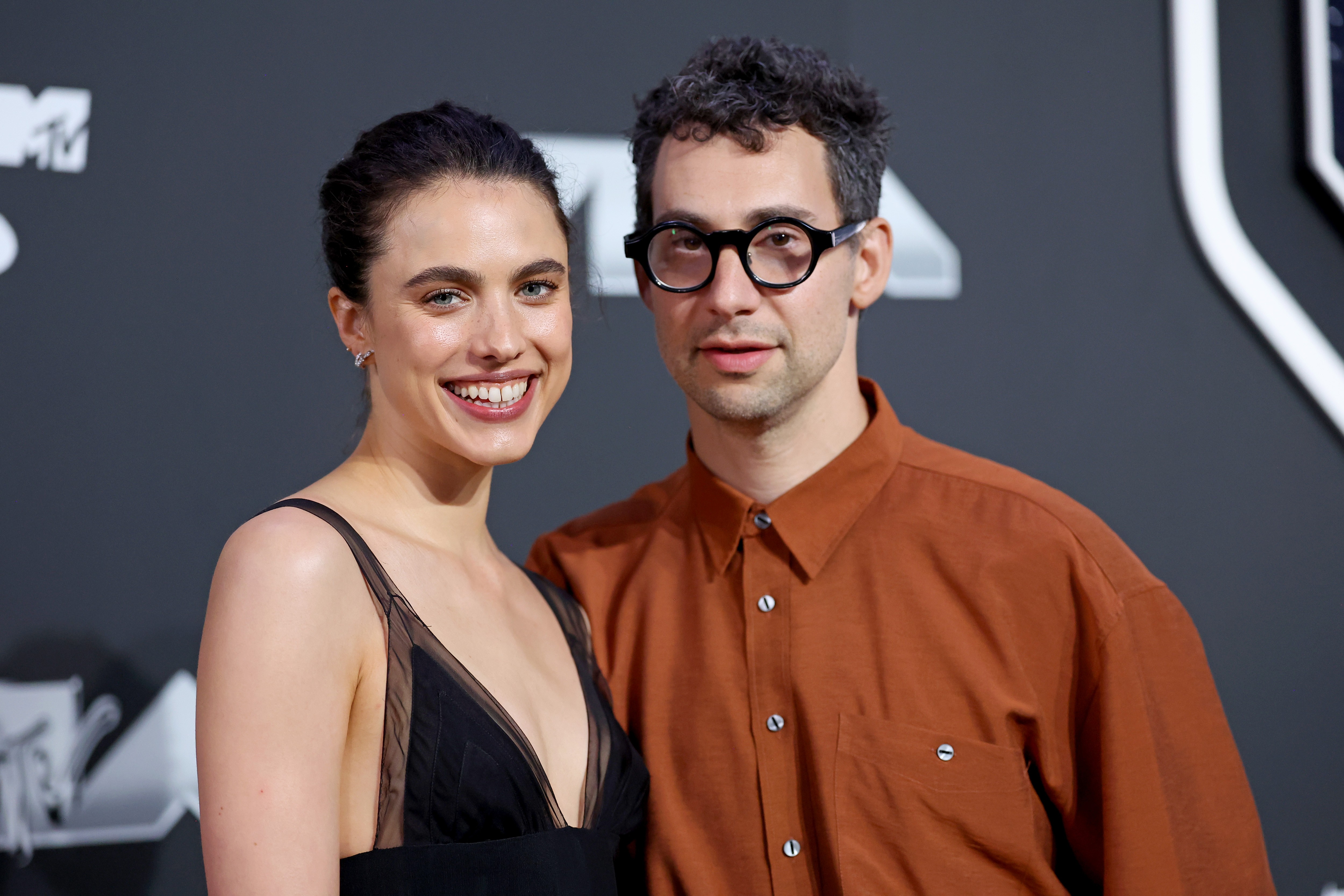 Margaret Qualley, sorrindo, com um vestido preto de alças finas e decote em V, ao lado de Jack Antonoff, que usa óculos pretos e camisa marrom, em um evento com fundo escuro e logo da MTV