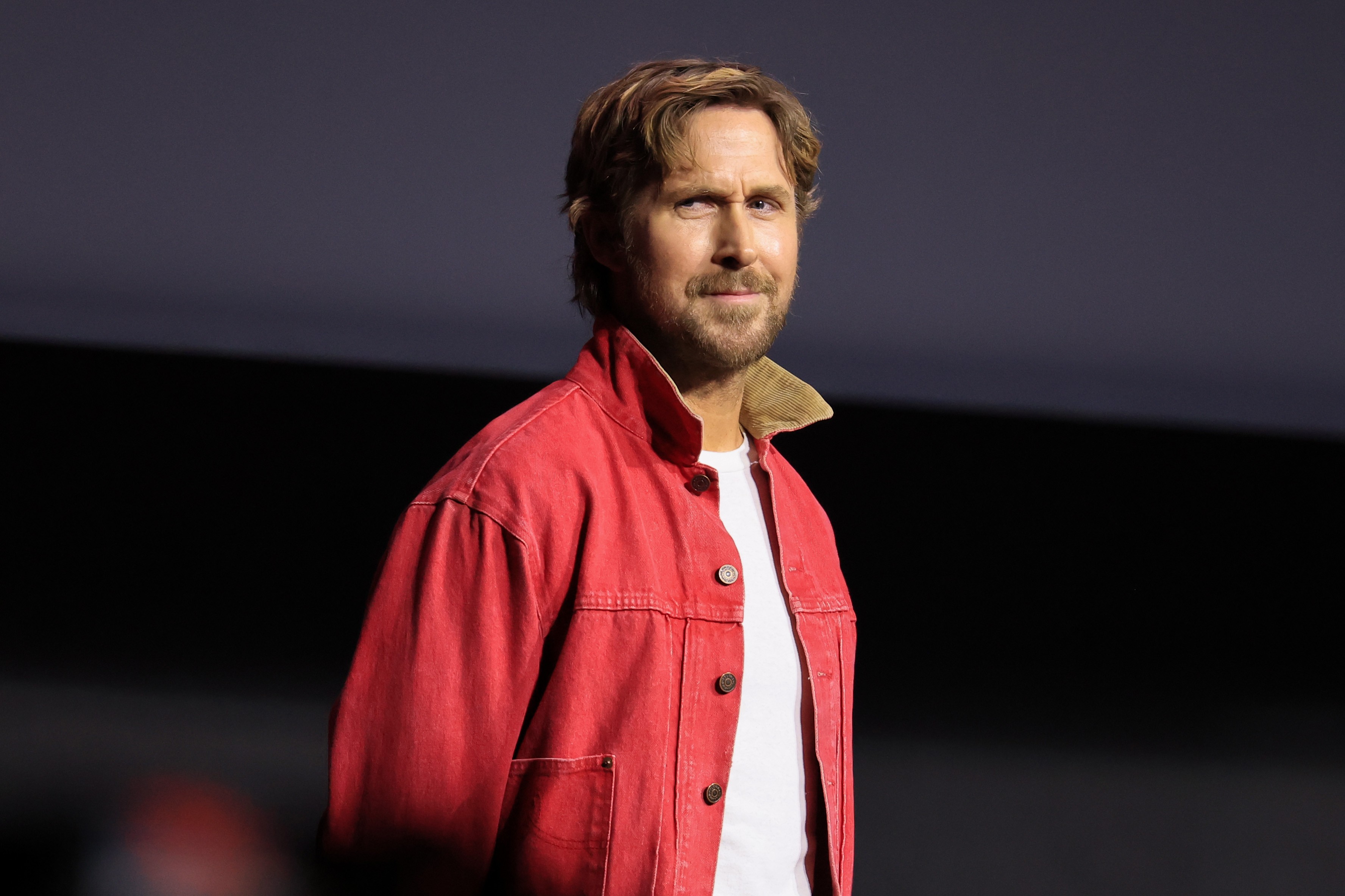 Ryan Gosling, ator de cabelo loiro-escuro e barba, veste jaqueta jeans vermelha com gola bege e camiseta branca, olhando para a frente com expressão séria em um fundo escuro
