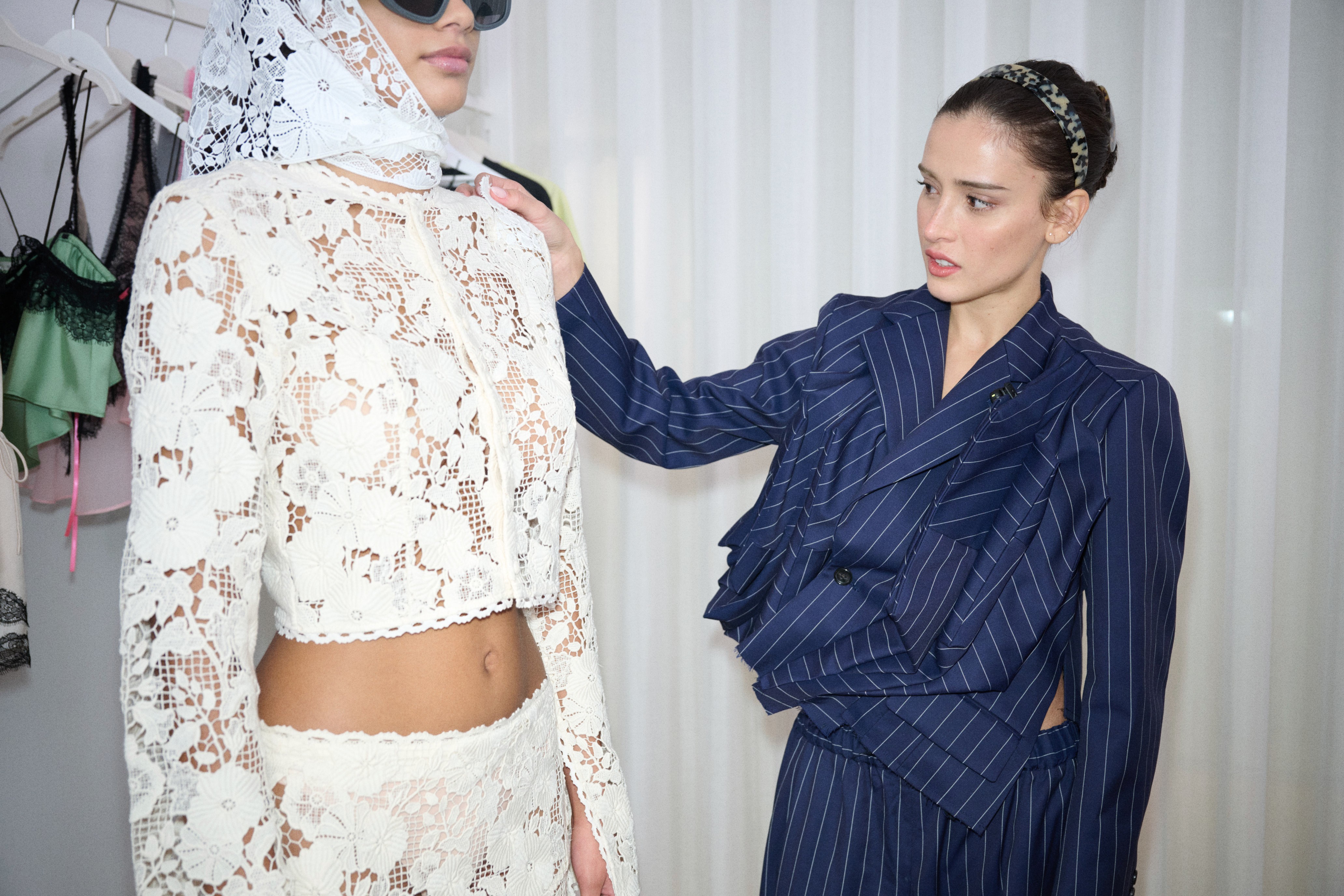 Duas mulheres em um provador. À esquerda, uma modelo de óculos escuros e lenço de renda branco na cabeça, vestindo top cropped e saia de renda creme. À direita, uma estilista de blazer e calça azul-marinho listrados, com tiara de oncinha, ajusta a roupa da modelo