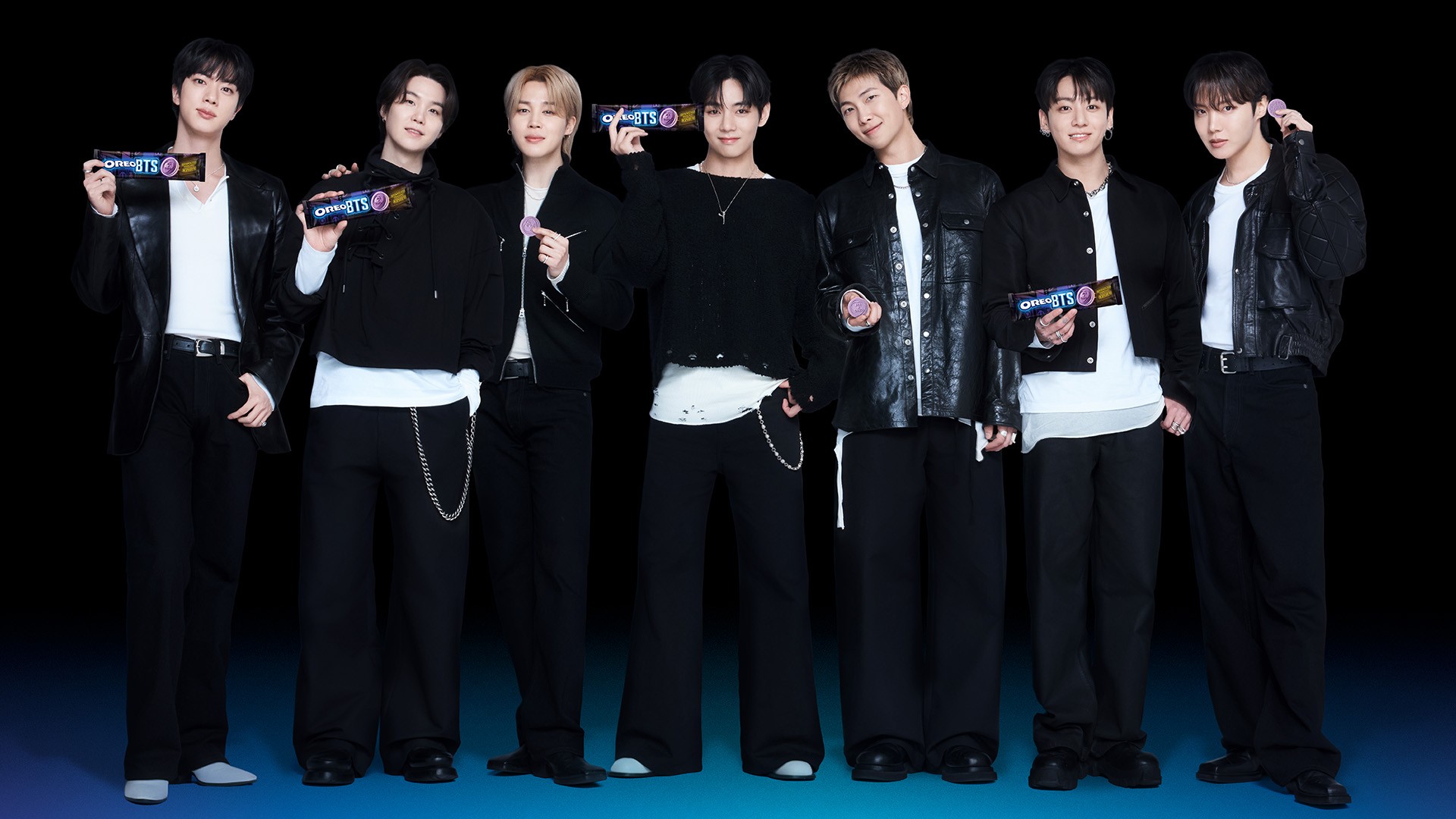 Sete homens jovens, membros do grupo BTS, posam sorrindo em fila, vestindo roupas pretas e brancas. Alguns seguram embalagens de chocolate roxas e pretas com o logo BTS, outros seguram pequenos objetos redondos roxos. O fundo é preto com uma faixa azul na parte inferior