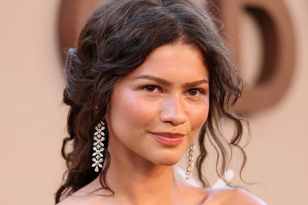 Zendaya, com cabelo castanho escuro e ondulado, usa brincos longos e brilhantes, sorrindo levemente para a câmera