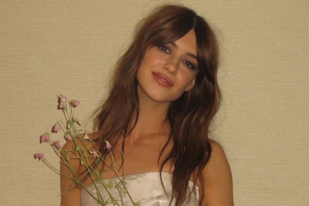 Mulher jovem de cabelos castanhos longos e franja, sorrindo levemente, segurando um pequeno buquê de flores cor-de-rosa, vestindo um tomara que caia branco, contra uma parede bege texturizada