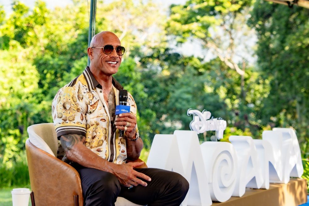 Dwayne The Rock Johnson sorri, usando óculos escuros e camisa estampada, sentado em uma cadeira marrom, segurando um microfone. Ao fundo, árvores e letras brancas formando MOANA com o logo da Disney