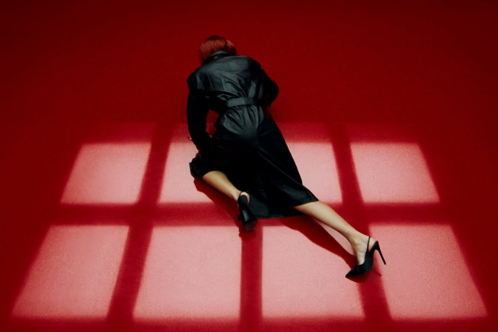 Mulher de cabelos ruivos e casaco preto, deitada de bruços no chão vermelho, com pernas esticadas e sapatos de salto alto. Luzes em formato de grade projetam sombras no chão.