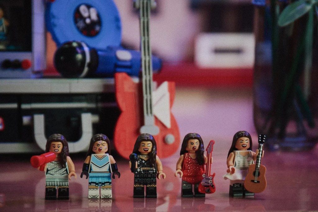 Cinco bonecas Lego femininas, com cabelos longos e castanhos, vestindo roupas de palco e segurando instrumentos musicais, como microfone, guitarra elétrica e violão, em um cenário de show com uma guitarra vermelha grande ao fundo