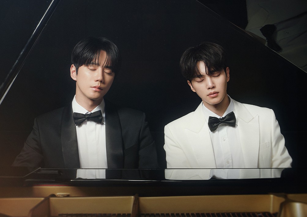 Dois homens jovens, um de terno preto e outro de terno branco, ambos com gravata borboleta preta, sentados ao piano de cauda com os olhos fechados, em um fundo escuro