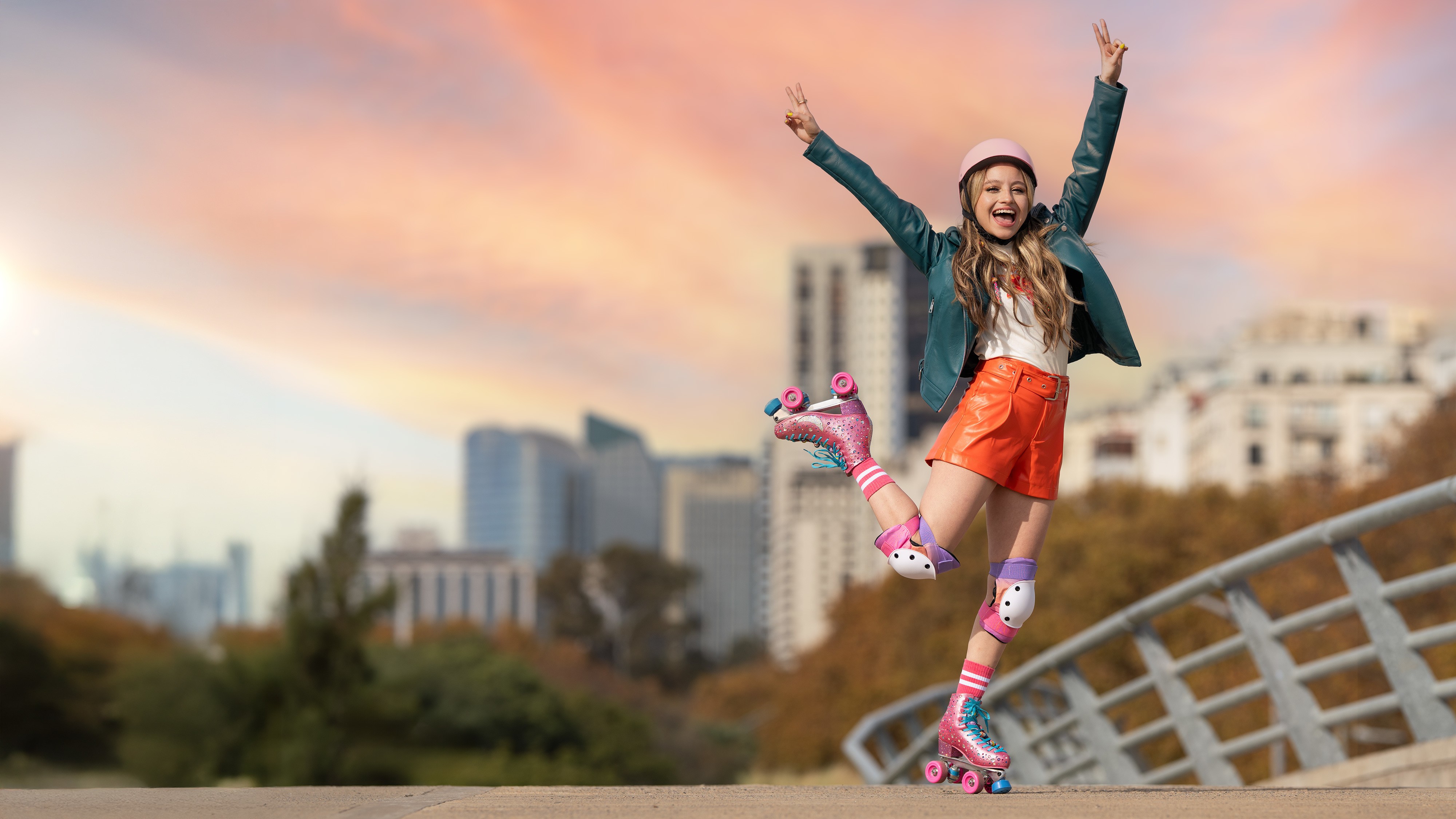 Jovem sorridente com capacete rosa e jaqueta verde, shorts laranja e patins brilhantes, braços erguidos em V, em ponte com cidade ao fundo e céu alaranjado