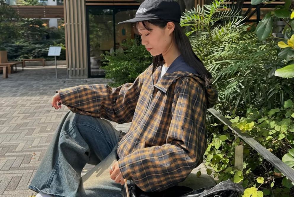 Jovem mulher asiática sentada ao ar livre, usando boné preto, jaqueta xadrez marrom e azul, calça jeans larga e tênis branco. Ela tem cabelos longos e escuros, e está olhando para baixo, com expressão pensativa. O fundo mostra um parque com árvores e arbustos verdes, e um edifício moderno com fachada de madeira. A luz do sol ilumina a cena, criando um clima tranquilo.