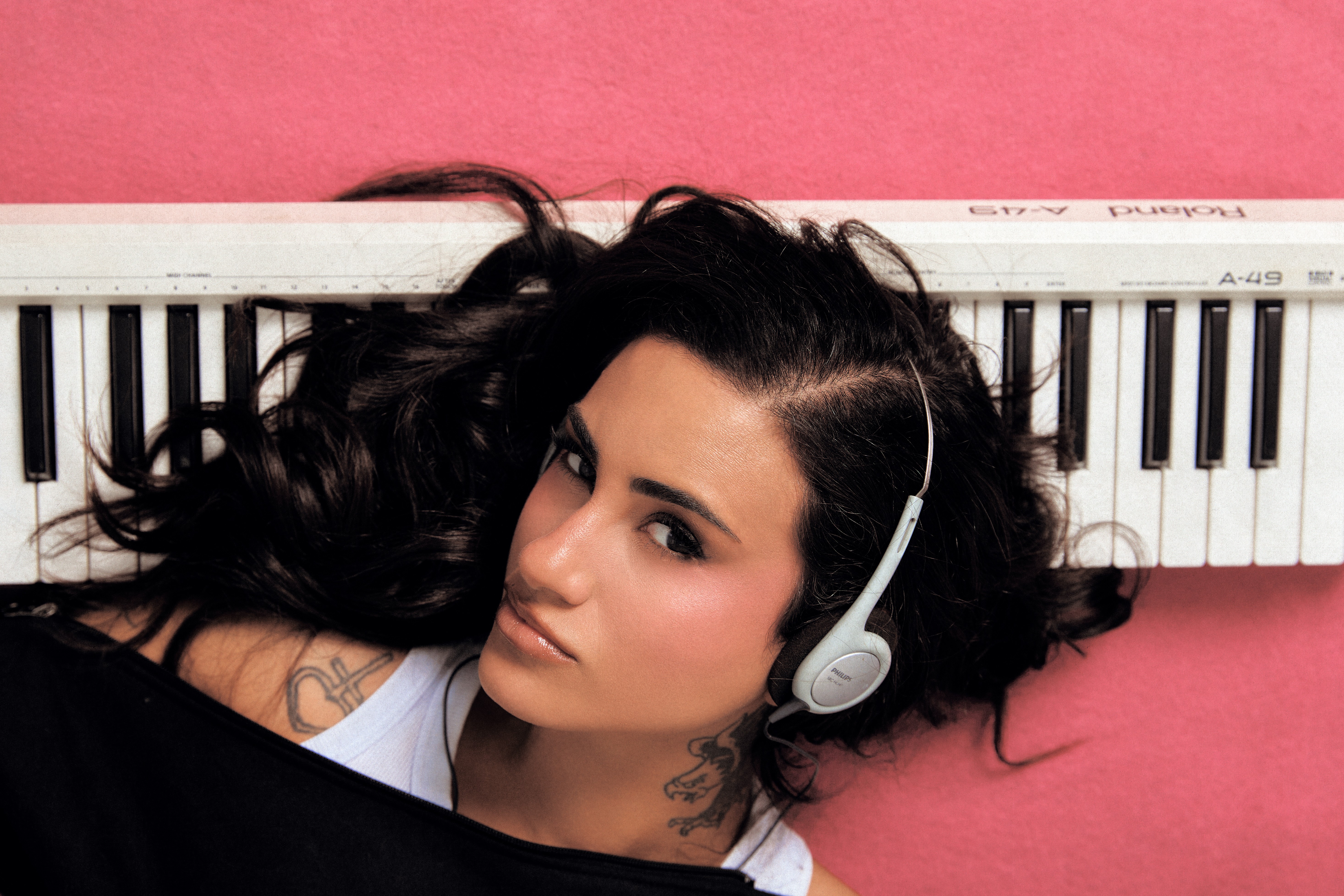 Mulher deitada de costas em um fundo rosa, com cabelos escuros espalhados e um teclado musical branco Roland A-49 acima da cabeça. Ela usa fones de ouvido cinza claro, uma camiseta branca e uma blusa preta, com tatuagens visíveis no pescoço e ombro. Seu olhar é direto para a câmera, com maquiagem leve e lábios brilhantes
