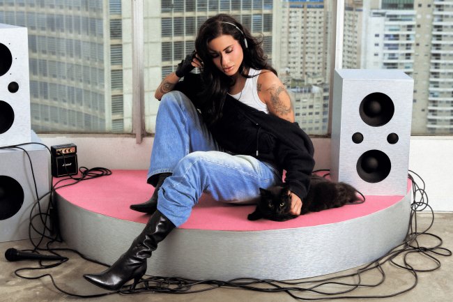 DAY LIMNS Mulher de cabelos escuros, com tatuagens nos braços, sentada em uma plataforma rosa e cinza, usando fones de ouvido, calça jeans larga, blusa branca, jaqueta preta e botas de cano alto. Ela acaricia um gato preto deitado ao seu lado. Ao redor, há caixas de som prateadas, um microfone e fios espalhados. Ao fundo, uma janela com prédios altos da cidade