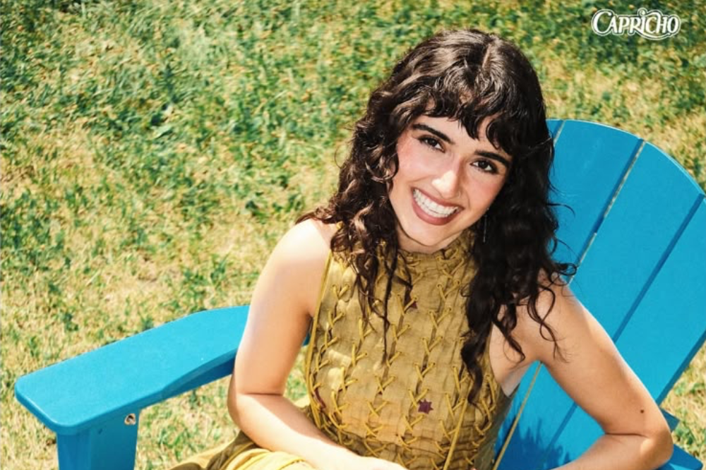 Jovem mulher de cabelos cacheados escuros e franja, sorrindo amplamente, sentada em uma cadeira azul de jardim, com gramado verde ao fundo