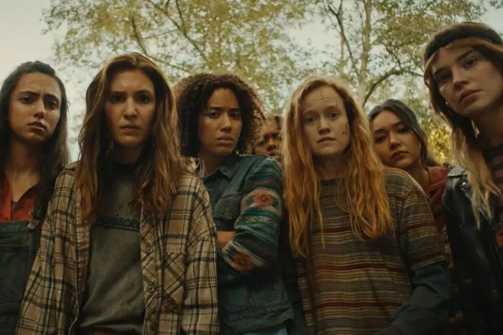 Um grupo de sete mulheres jovens, com expressões sérias e olhares fixos, em um cenário externo com árvores ao fundo. Elas vestem roupas casuais e algumas têm o rosto sujo ou com pequenos arranhões, sugerindo uma situação de sobrevivência ou conflito
