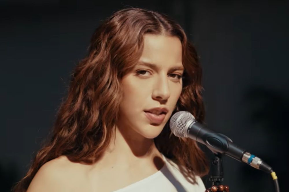 Jovem mulher de cabelos castanhos ondulados, vestindo blusa branca de um ombro só, cantando em um microfone prateado com fundo escuro