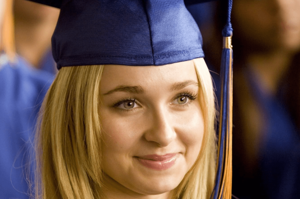 Jovem mulher loira, com olhos claros e maquiagem leve, sorri suavemente enquanto usa um capelo de formatura azul com um pendão azul e dourado. Ela está em um ambiente interno, possivelmente uma cerimônia de graduação, com outras pessoas em segundo plano.