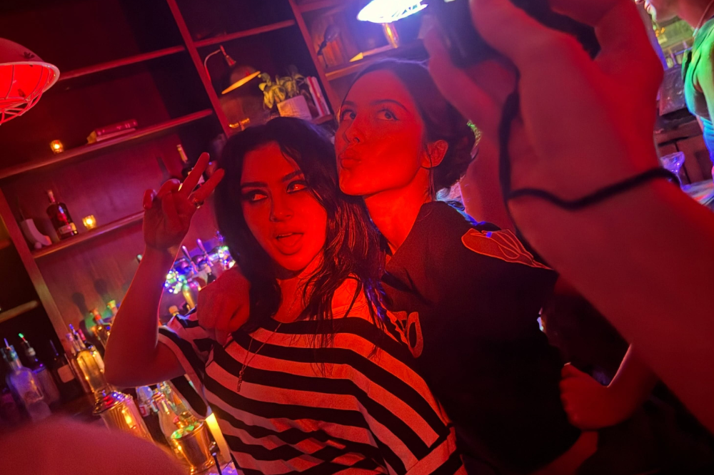 Duas mulheres posam para selfie em bar com iluminação vermelha. A da frente, de blusa listrada, faz sinal de paz com a mão e mostra a língua. A de trás, de blusa escura, faz bico. Garrafas e copos coloridos no fundo