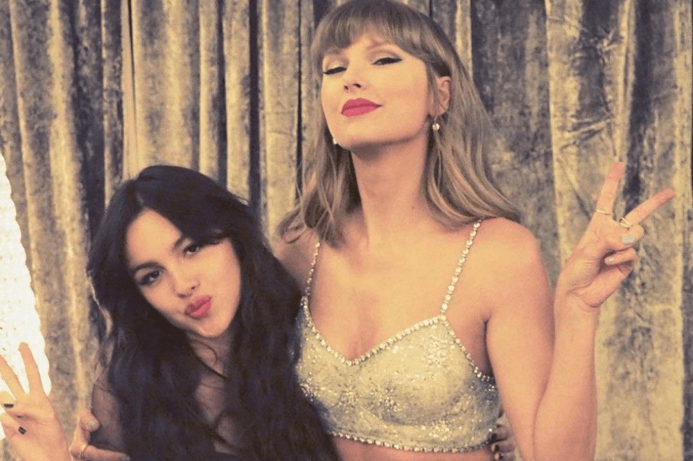 Taylor Swift, loira de franja e batom vermelho, usa top brilhante e faz sinal de paz. Olivia Rodrigo, morena de batom rosa, faz bico e sinal de paz, abraçando Swift. Fundo de cortina dourada.