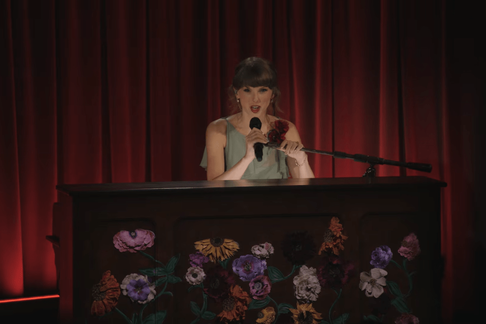 Taylor Swift, com um vestido azul claro, canta em um microfone enquanto segura uma rosa vermelha, atrás de um piano escuro decorado com flores coloridas, em um palco com cortinas vermelhas