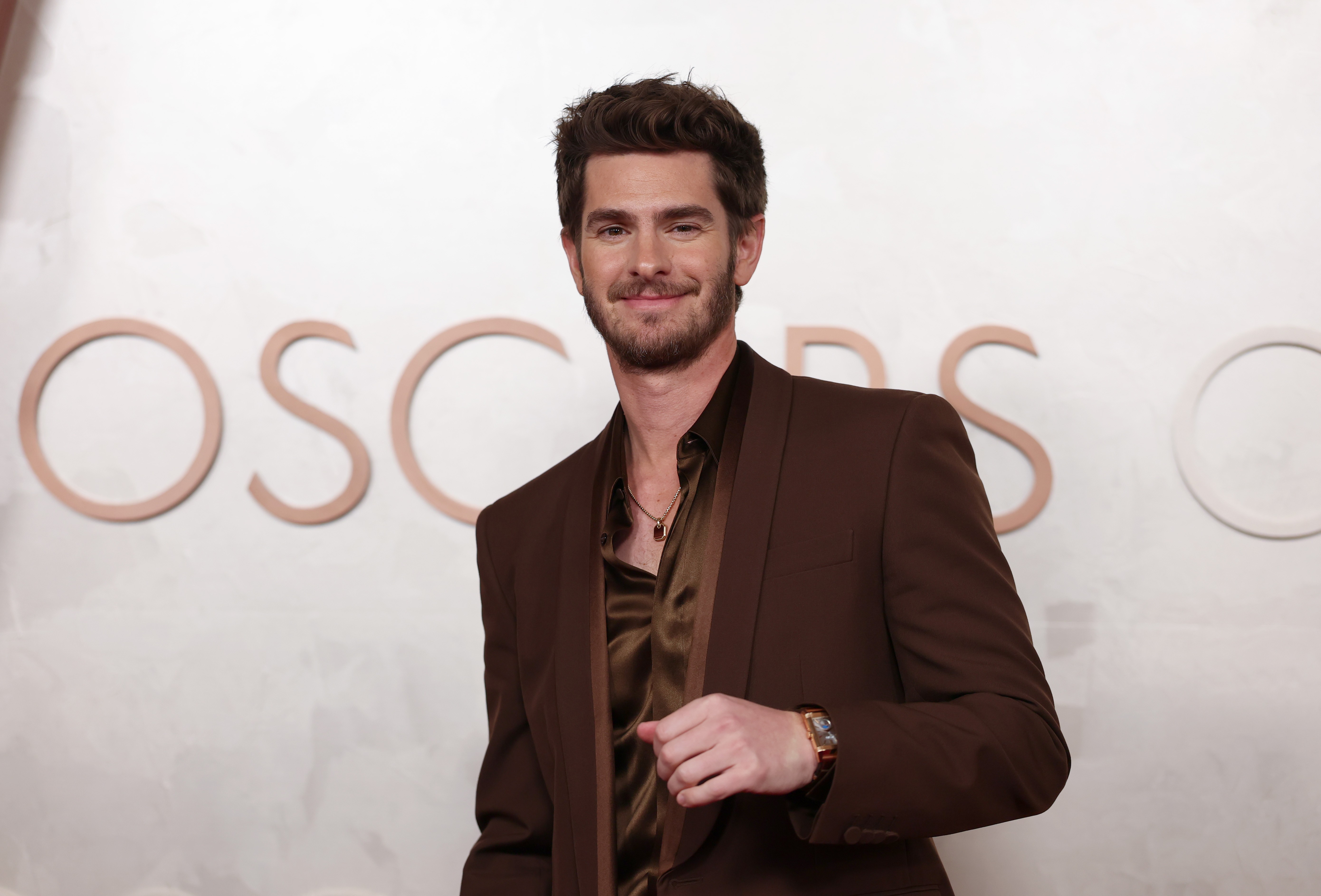 Andrew Garfield sorrindo, com barba e cabelo castanho, vestindo um terno marrom e camisa de seda, com um colar e relógio. Ao fundo, letras grandes e claras formam OSCARS