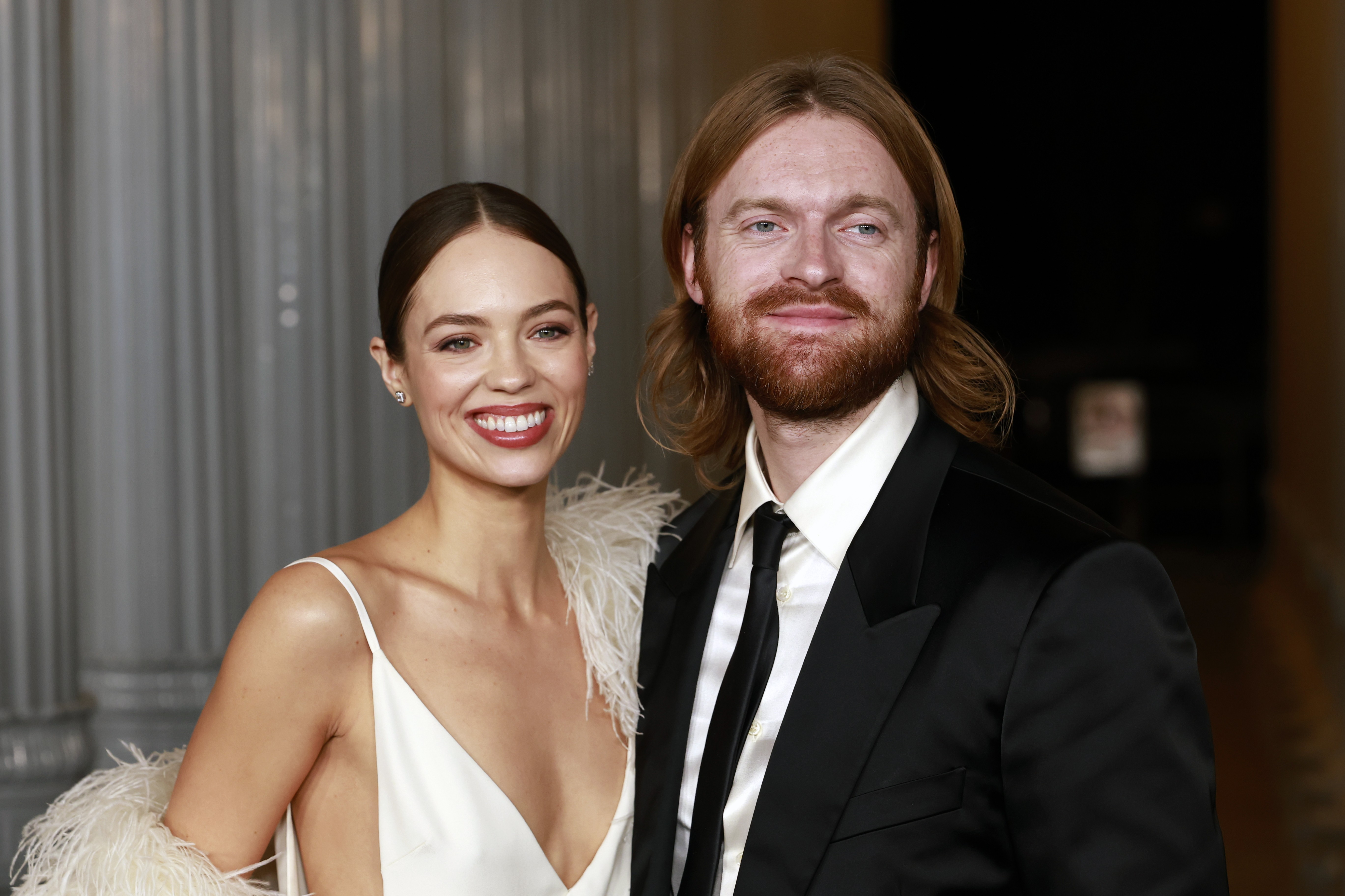 Uma mulher sorridente com vestido branco de alças finas e estola de penas claras ao lado de um homem barbudo de cabelos longos e terno preto com gravata, ambos em evento formal