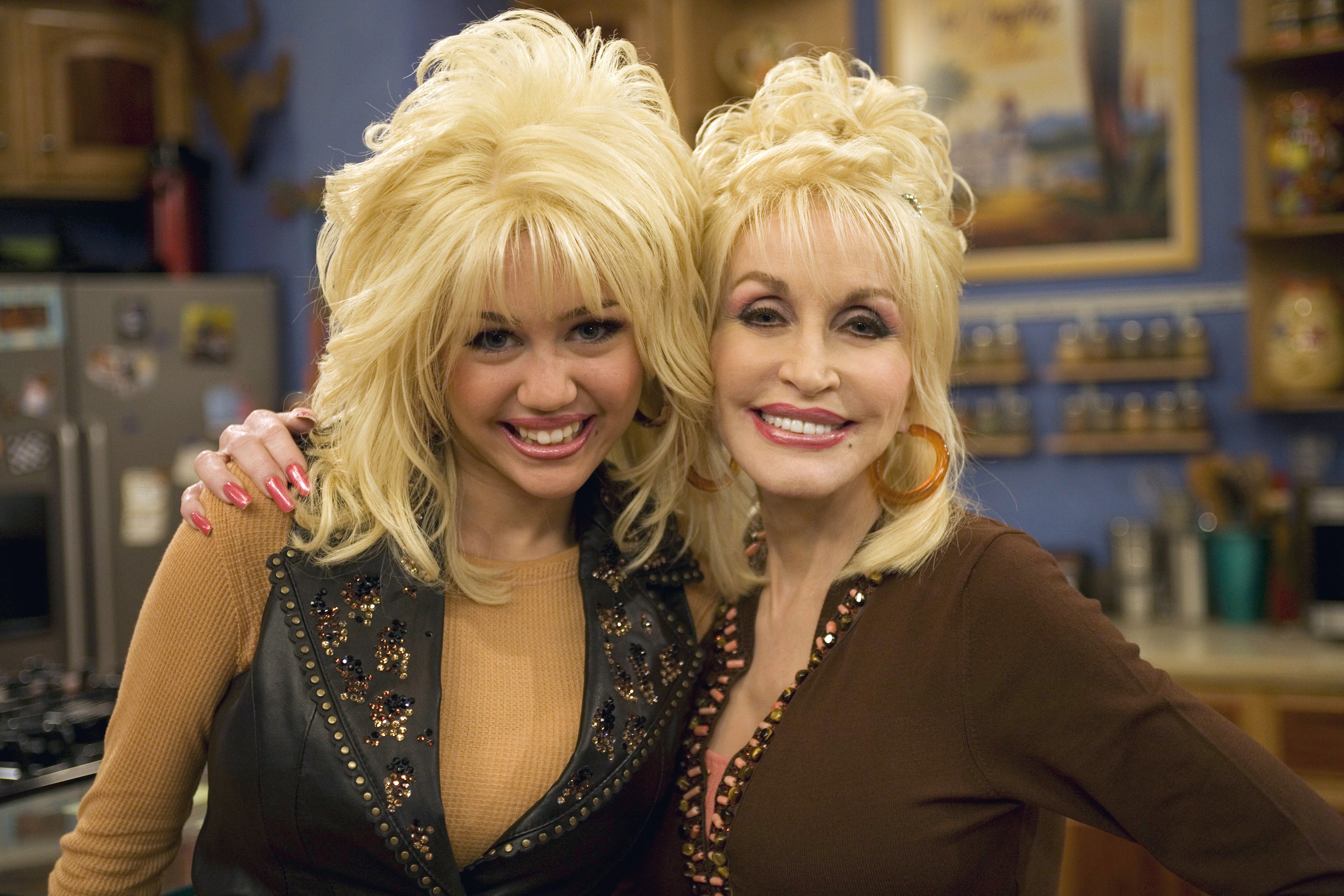 Miley Cyrus e Dolly Parton sorrindo para a câmera, ambas com perucas loiras volumosas, maquiagem marcante e roupas escuras com brilhos, em um cenário de cozinha