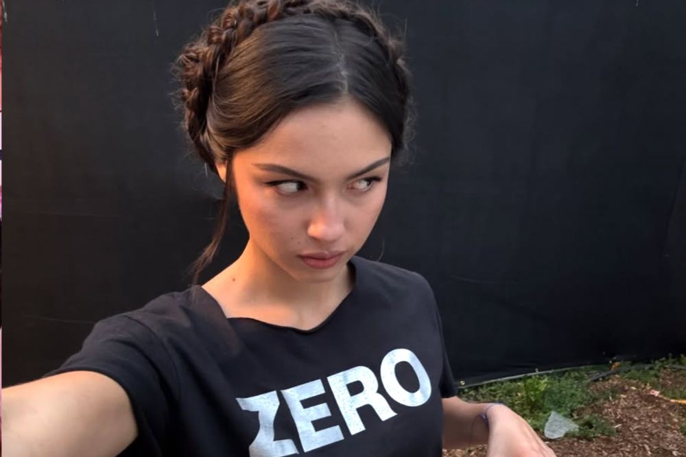 Mulher jovem com cabelo escuro preso em tranças, vestindo camiseta preta com a palavra ZERO em branco, olhando para o lado com expressão séria, em fundo escuro e grama