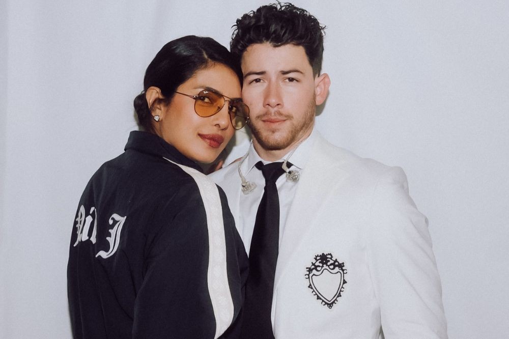 Priyanka Chopra, de óculos de sol laranja e jaqueta preta com listra branca, abraça Nick Jonas, de terno branco e gravata preta, com fones de ouvido e um brasão bordado no paletó