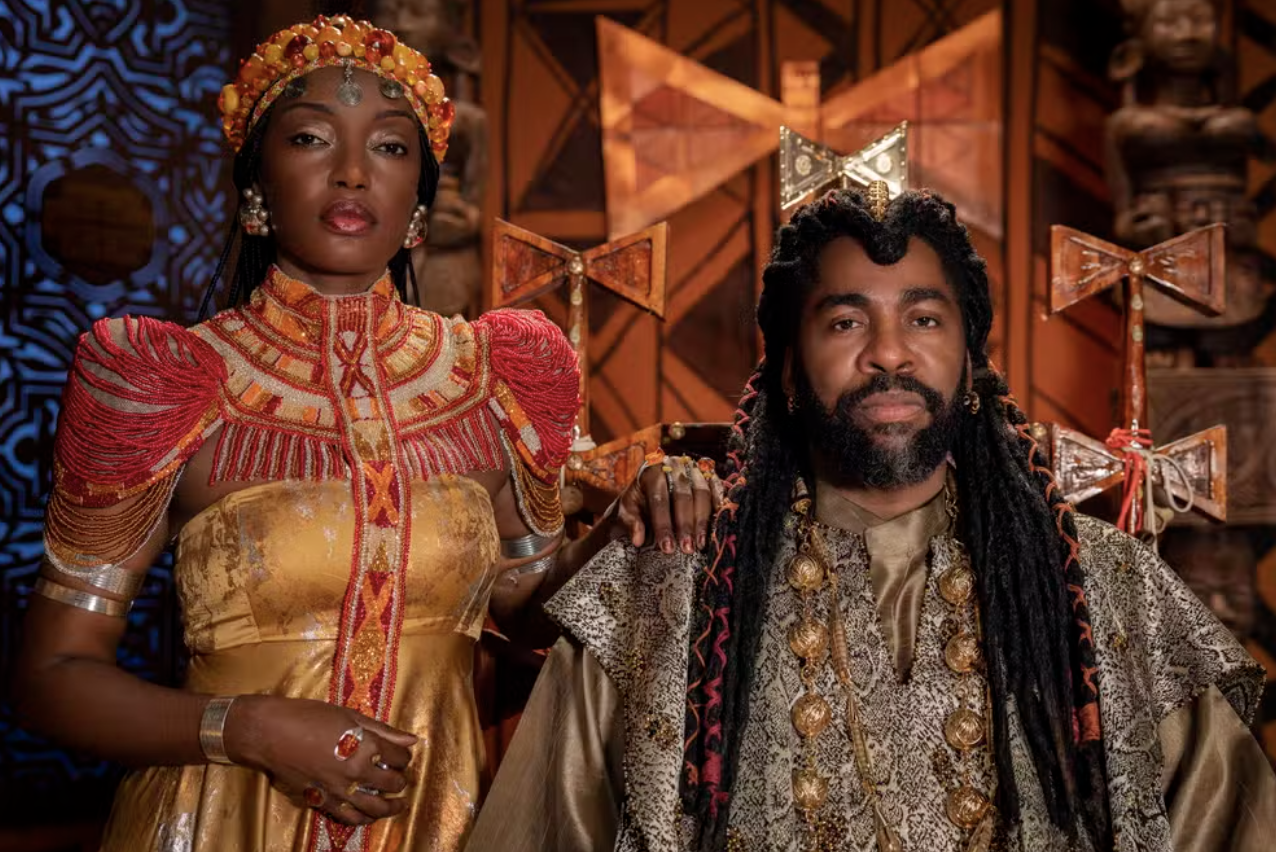 Uma mulher e um homem negros, vestidos com trajes cerimoniais africanos, posam com expressões sérias. Ela usa um vestido dourado com ombros e gola adornados por miçangas vermelhas e douradas, e uma coroa de miçangas. Ele tem dreadlocks longos, barba e veste uma túnica estampada com colares dourados. Ao fundo, elementos decorativos em madeira escura e dourada.