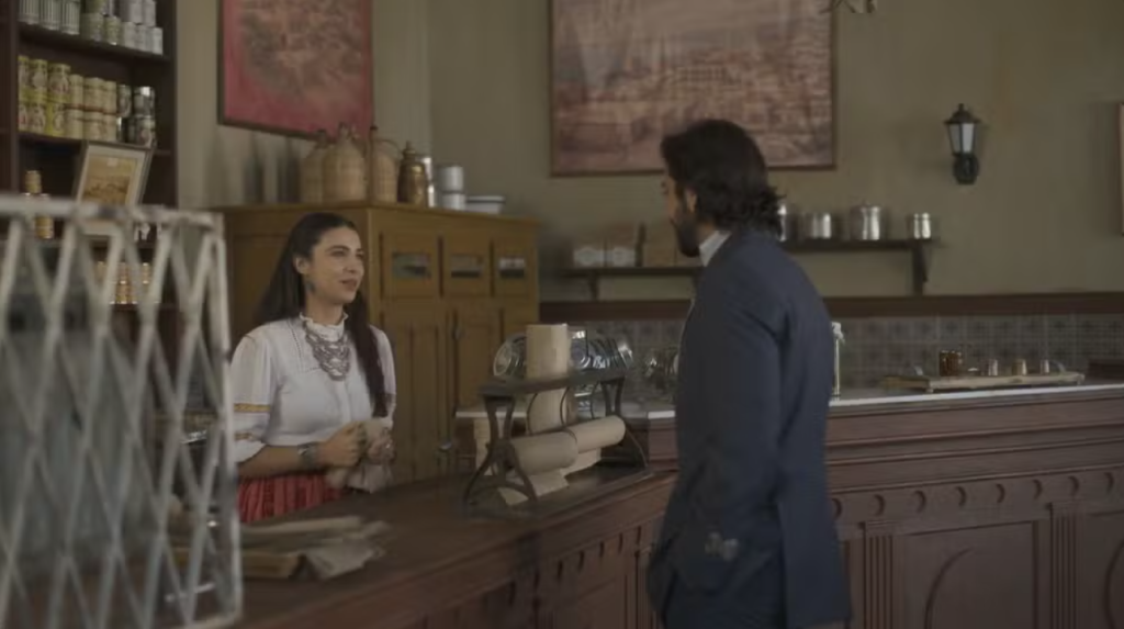 Mulher sorridente com blusa branca e colar grande atende um homem de costas, com barba e casaco azul, em um balcão de madeira de uma loja antiga com prateleiras cheias de potes e quadros na parede