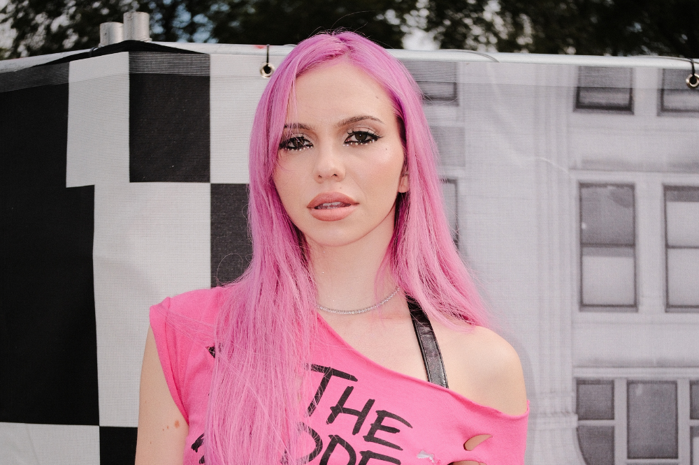 Mulher jovem com cabelo rosa vibrante e maquiagem escura nos olhos, vestindo uma camiseta rosa rasgada e colar brilhante, posa séria em frente a um fundo quadriculado preto e branco e um prédio cinza