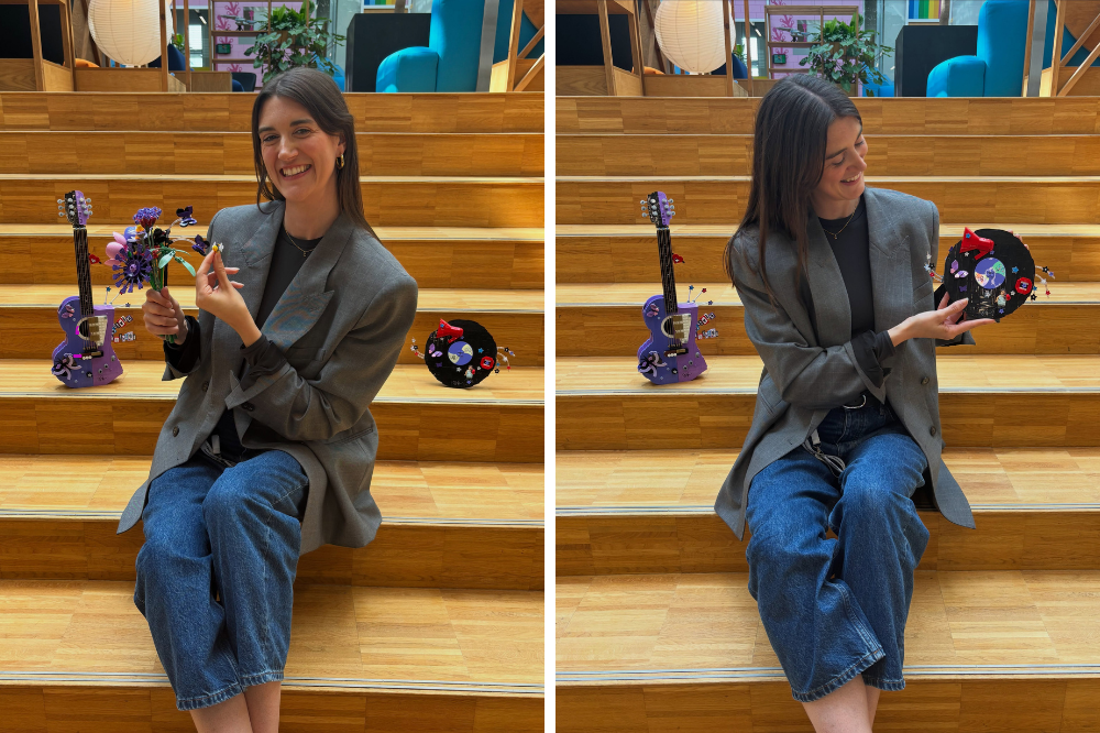 Mulher sorridente sentada em escada de madeira, vestindo blazer cinza e jeans, segurando um buquê de flores roxas e amarelas de Lego na mão esquerda e um pequeno elemento amarelo na direita. Ao lado, uma guitarra roxa e um disco de vinil preto, ambos decorados com peças de Lego coloridas. Na imagem à direita, ela aponta para o disco de vinil, sorrindo