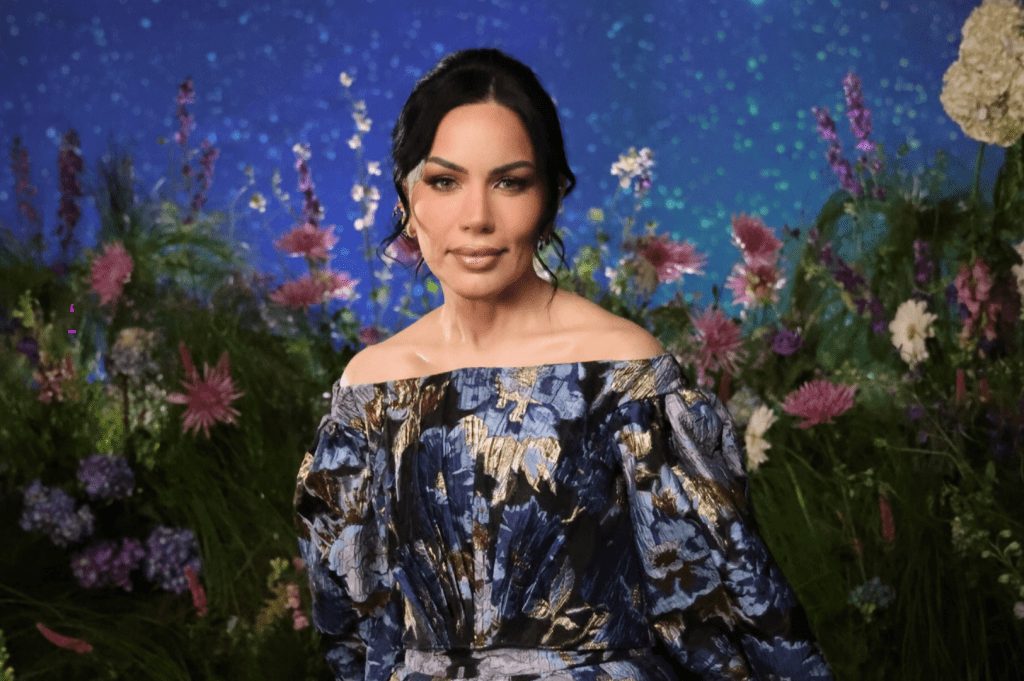 Mulher de pele clara, cabelo escuro preso com mechas brancas na frente, maquiagem leve, usando um vestido azul e dourado com ombros à mostra, posa sorrindo em um cenário de jardim com flores coloridas e fundo azul estrelado