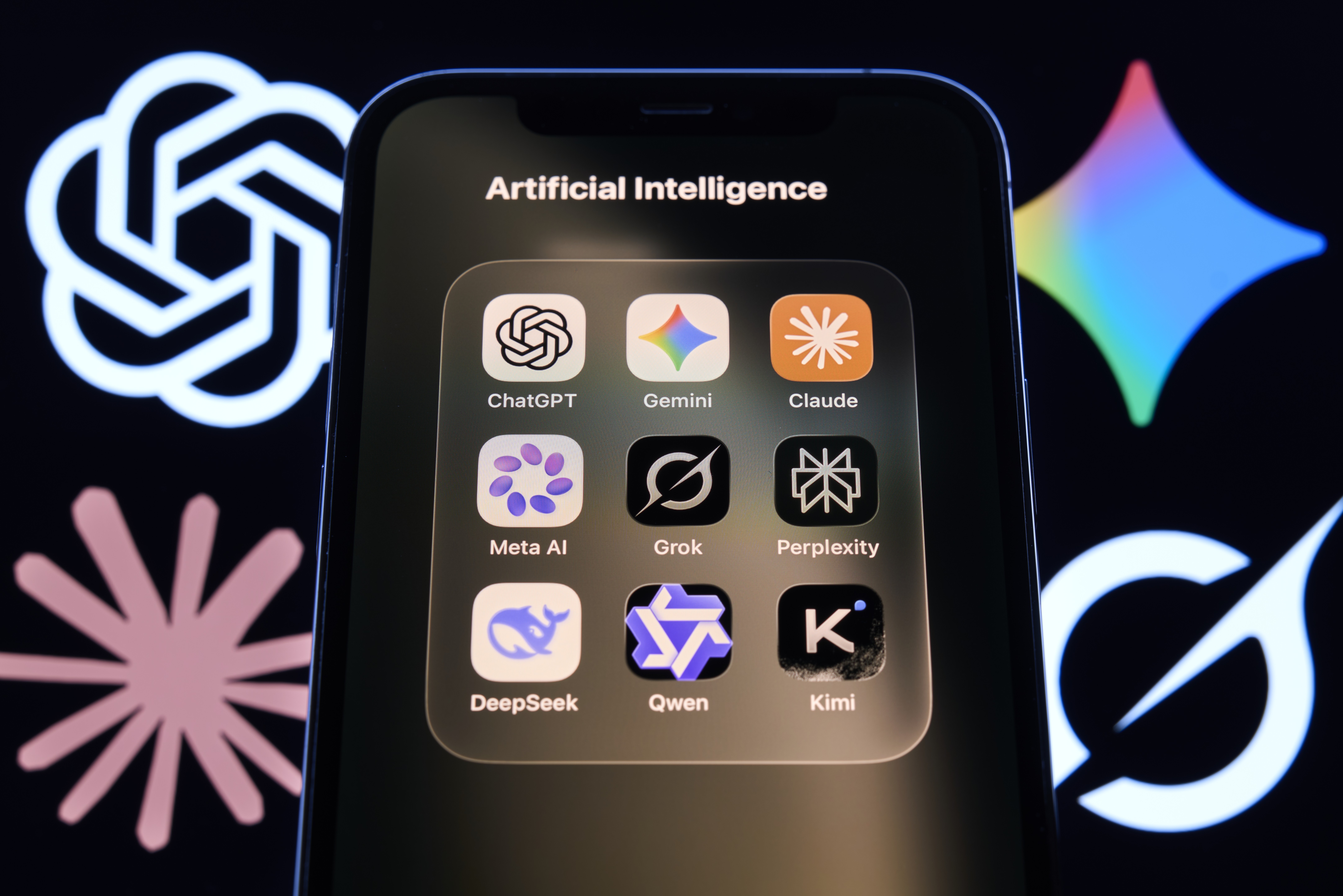 Tela de smartphone exibindo uma pasta de aplicativos chamada Artificial Intelligence, com ícones de ChatGPT, Gemini, Claude, Meta AI, Grok, Perplexity, DeepSeek, Qwen e Kimi. Ao fundo, logotipos de IA em branco e colorido em um monitor escuro