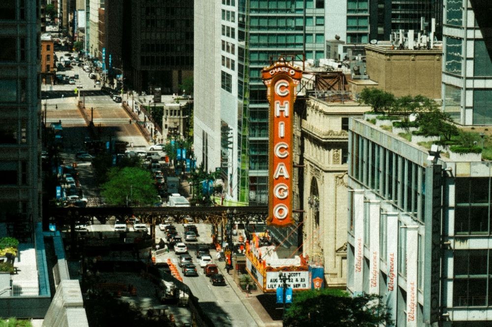 Vista aérea da rua State Street em Chicago, com o letreiro vertical vermelho do Teatro Chicago em destaque. Carros e ônibus trafegam pela rua, ladeada por edifícios altos e modernos. Um viaduto de trem elevado atravessa a imagem, e faixas da Walgreens são visíveis em um prédio à direita