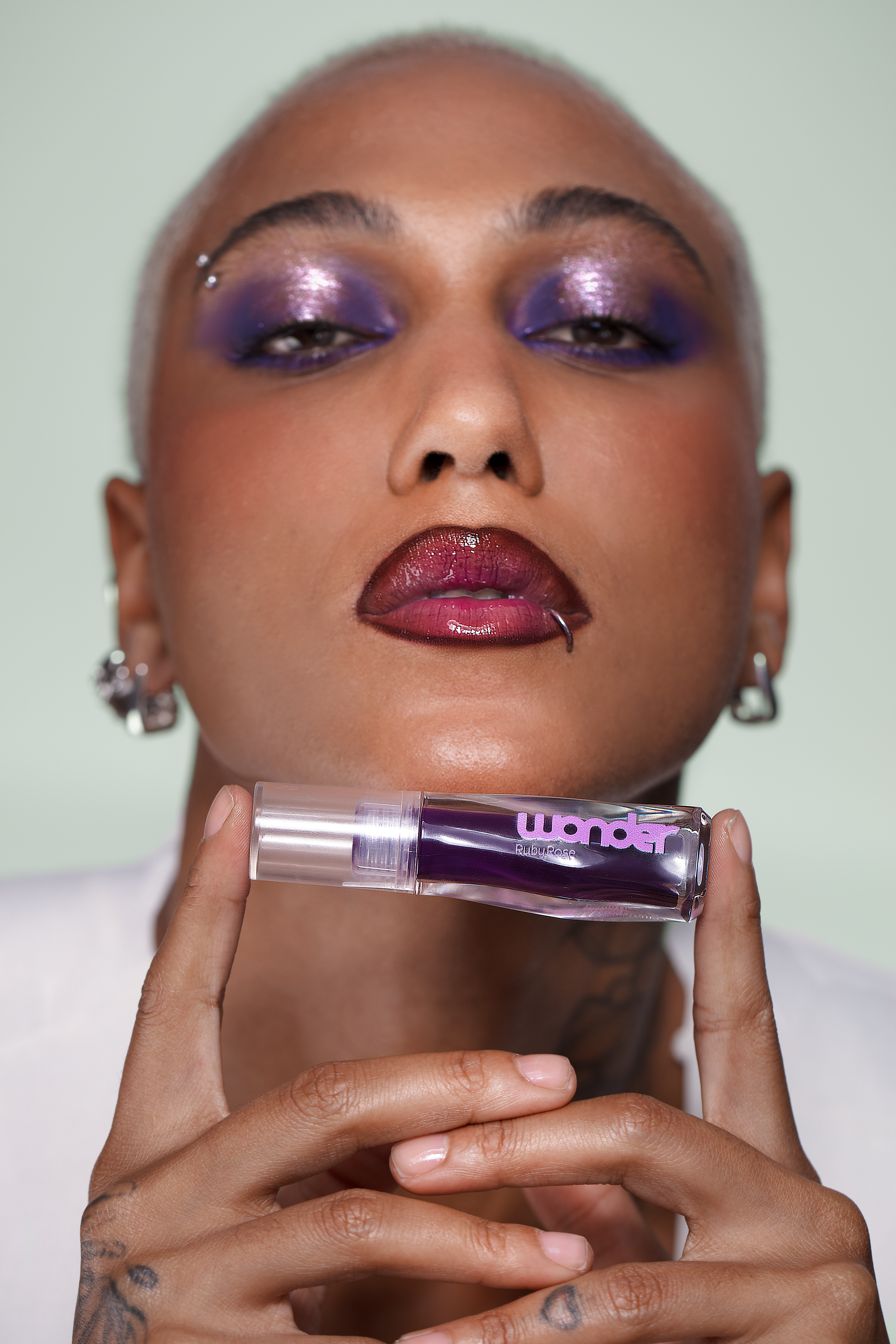 Pessoa de pele escura, cabeça raspada, maquiagem roxa brilhante nos olhos e batom vinho, segurando um gloss labial roxo da marca Wonder Ruby Rose com as duas mãos, contra um fundo verde claro