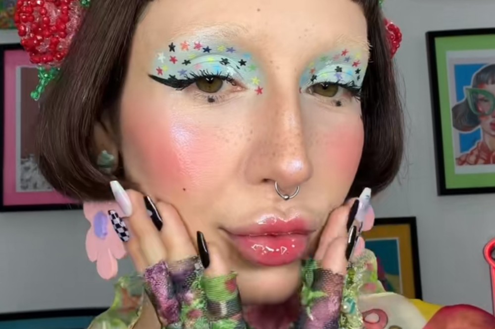 Close-up de uma pessoa com maquiagem vibrante, bochechas rosadas, lábios brilhantes e delineado preto com estrelas coloridas nas pálpebras. Ela usa um piercing no nariz, unhas longas pretas e brancas, e luvas de renda floral.