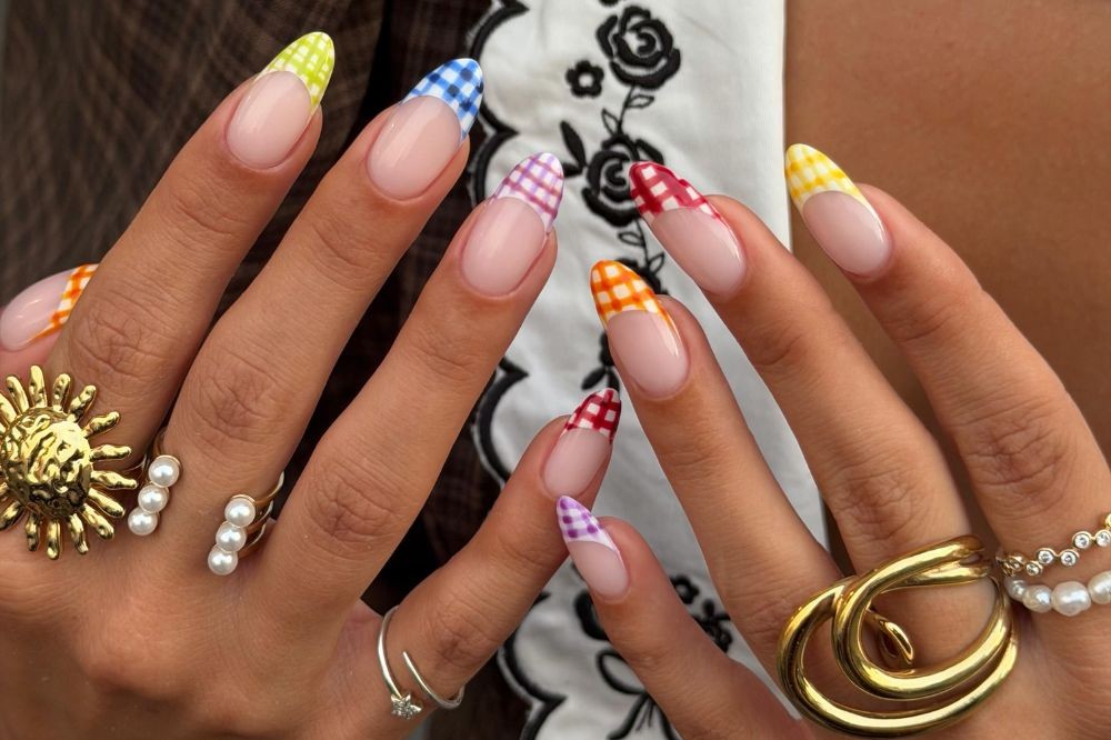 Mãos com unhas amendoadas e francesinha colorida em xadrez vichy: verde, azul, roxo, vermelho, laranja e amarelo. A mão esquerda exibe anéis dourados de sol e pérolas, e um anel prateado com flor. A mão direita tem um anel dourado grande e um anel de pérolas