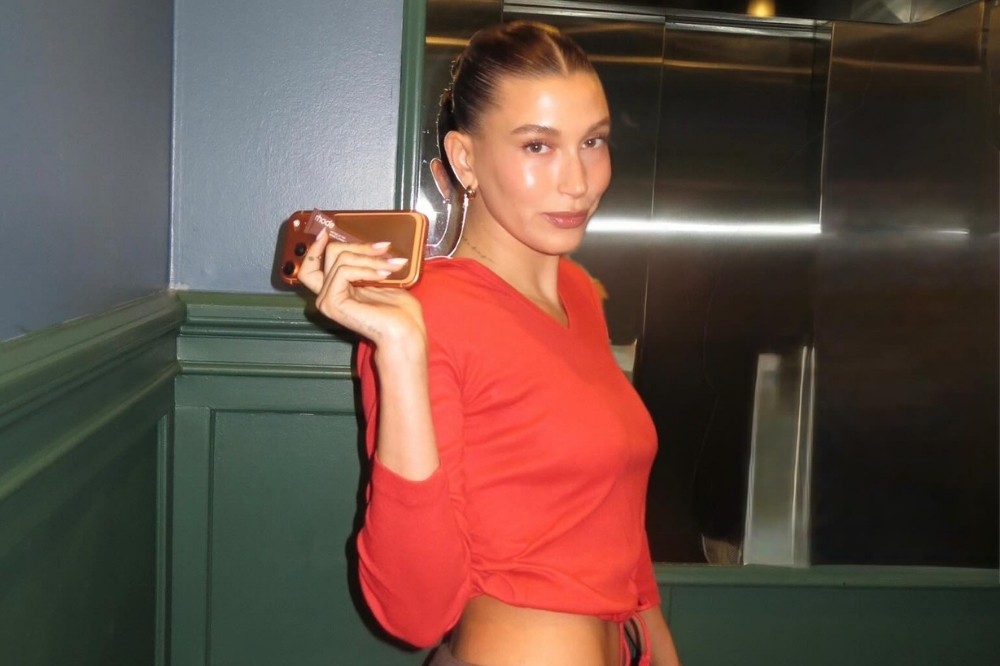 Mulher jovem com cabelo castanho preso, usando blusa laranja de manga comprida e calça marrom, posa sorrindo levemente enquanto segura um celular dourado na mão esquerda. Ela está em um ambiente com paredes azuis e rodapés verdes, ao lado de um elevador metálico