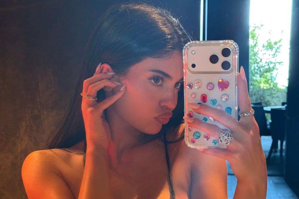 Jovem mulher de cabelos escuros e pele clara, com maquiagem leve, fazendo bico para uma selfie. Ela usa um top escuro, anéis e segura um celular com capa transparente decorada com pedras coloridas em formato de coração e redondo. A iluminação avermelhada realça seu rosto e ombro, com um fundo escuro e uma janela com folhagens verdes à direita