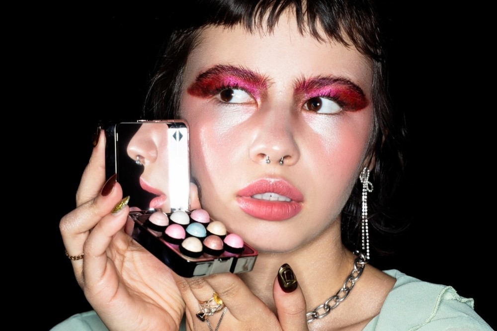Mulher jovem com maquiagem vibrante, sobrancelhas e pálpebras em tons de rosa e vermelho, segurando uma paleta de maquiagem com espelho que reflete seu nariz e boca. Ela tem piercing no septo, colar de corrente prateada e brincos longos, olhando para a direita com expressão pensativa, em fundo preto