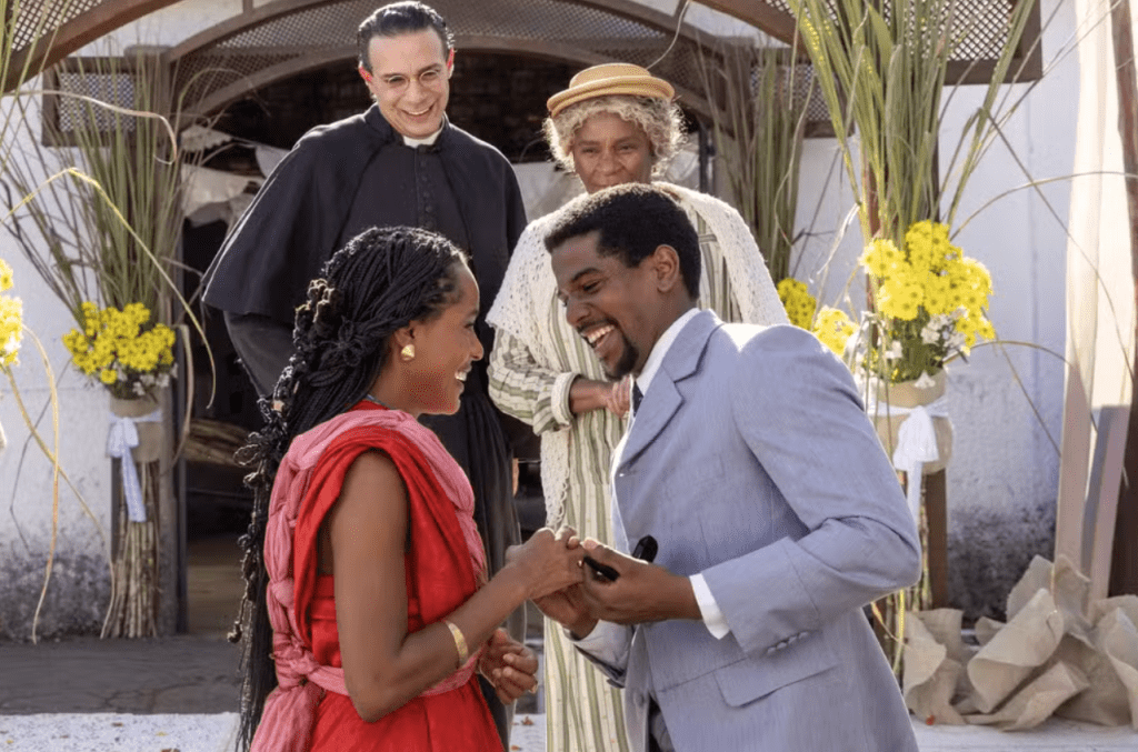 Um casal negro sorridente, ele de terno cinza claro e ela de vestido vermelho, trocando alianças. Ao fundo, um padre e uma mulher mais velha, todos felizes, em um cenário decorado com flores amarelas e folhagens, sugerindo uma cerimônia ao ar livre