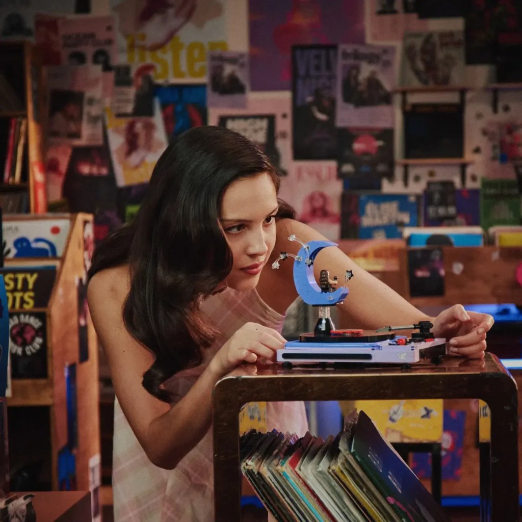Jovem mulher de cabelos escuros e ondulados, vestindo regata clara, observa atentamente um toca-discos de brinquedo com um enfeite de lua azul. Ela está em um quarto com paredes cobertas por pôsteres e prateleiras cheias de discos de vinil