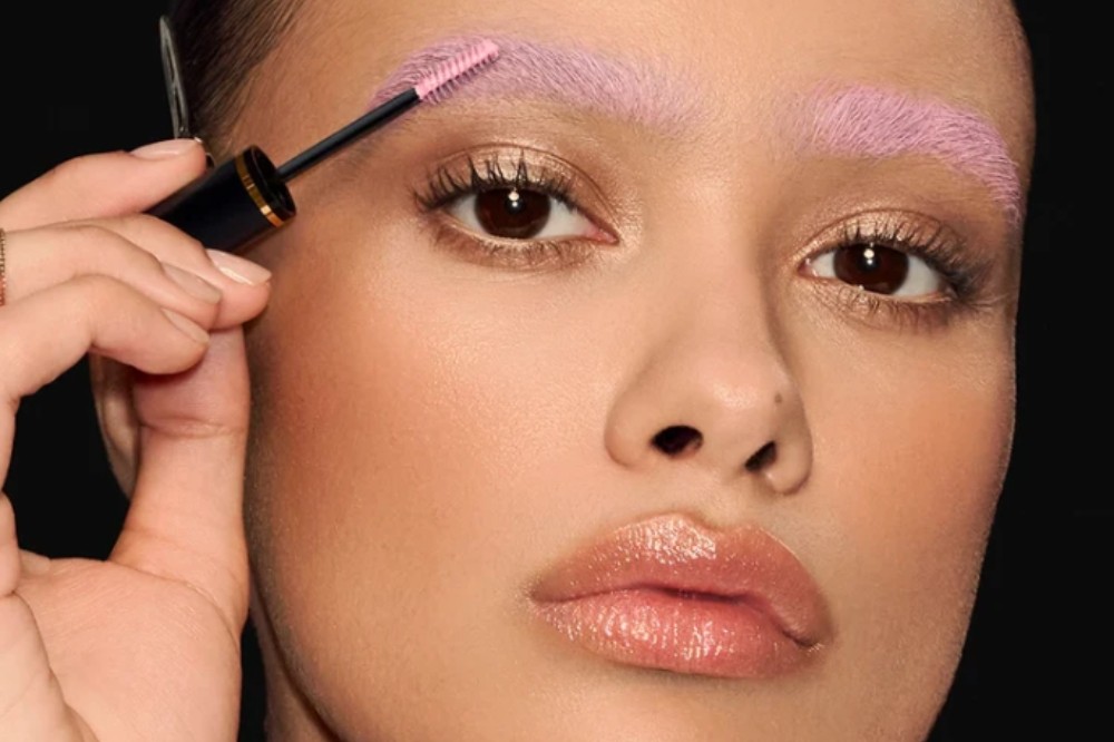 Close-up de uma mulher com pele morena aplicando rímel rosa nas sobrancelhas, que já estão tingidas de rosa claro. Ela tem maquiagem dourada nos olhos e lábios com gloss nude