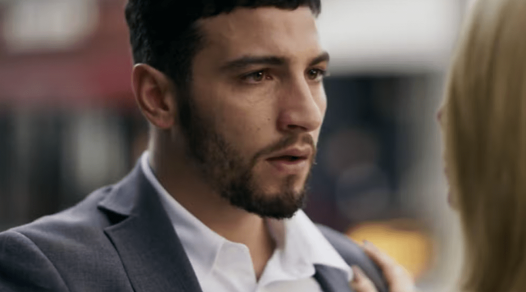 Homem de barba escura e cabelo curto, vestindo terno cinza e camisa branca, olha com expressão séria para uma mulher loira, cujo ombro direito e parte do cabelo são visíveis à direita