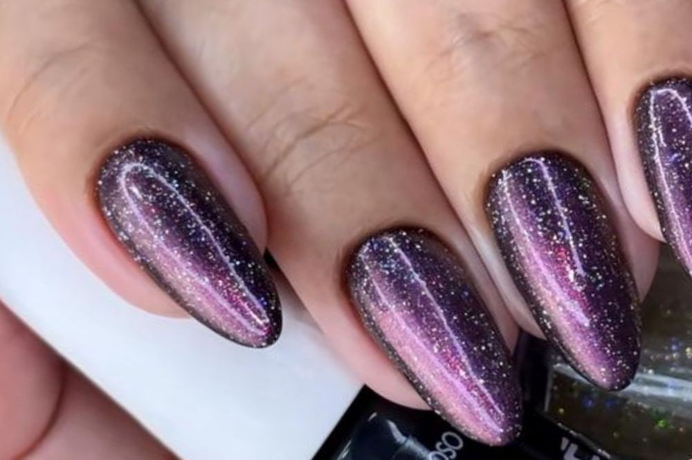 Mão feminina com unhas ovais longas pintadas de roxo escuro com brilho prateado e um efeito magnético rosa claro no centro