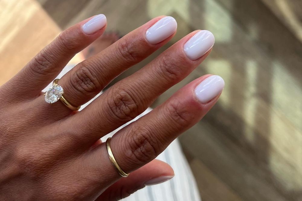 Mão de pele morena com unhas curtas pintadas de branco leitoso, usando um anel de ouro com diamante oval no dedo anelar e uma aliança fina de ouro no dedo médio
