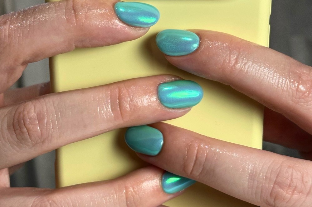 Mãos femininas segurando um celular amarelo, com unhas curtas pintadas de esmalte azul-turquesa iridescente, refletindo tons de verde e roxo