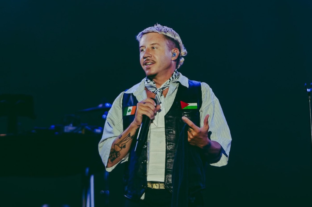 Homem branco de cabelo loiro platinado e bigode, vestindo colete preto sobre camisa listrada e lenço no pescoço, segurando um microfone e apontando para a bandeira da Palestina em seu colete, com a bandeira do México no outro lado. Ele está em um palco escuro, com iluminação azul ao fundo
